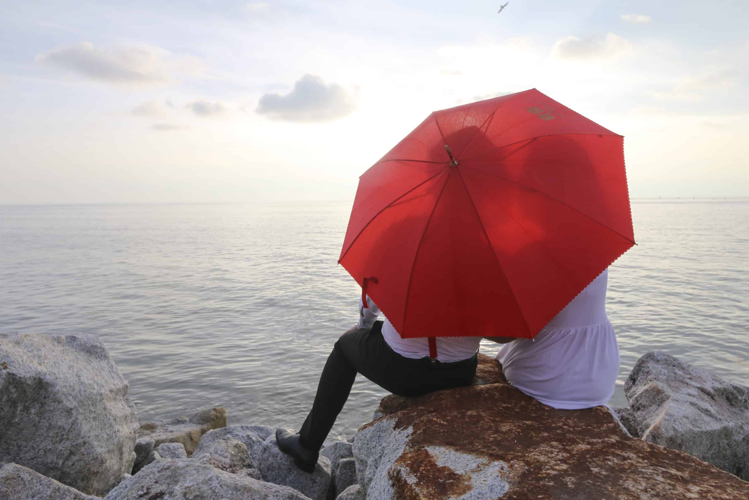 Paar sitzt gemeinsam geschützt unter einem roten Schirm am Meer